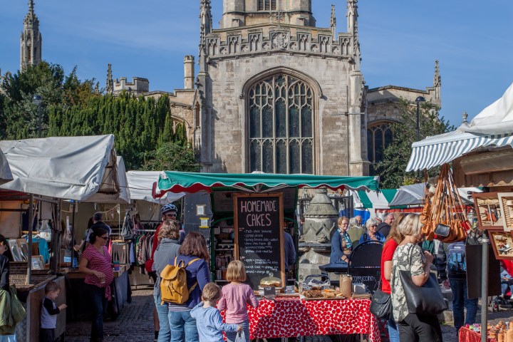 People at an outdoor market with homemade cakes, historic building in background.