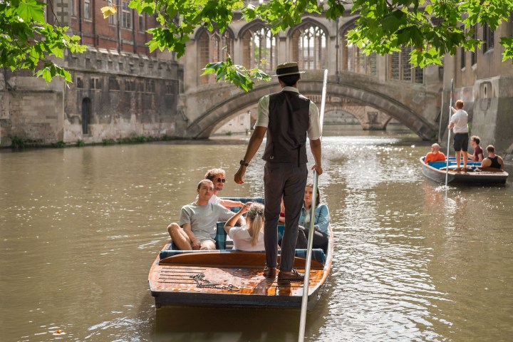 People enjoying a punt ride on a river under a historic stone bridge on a sunny day.