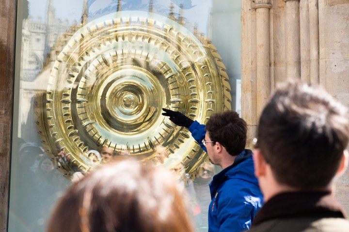 Man pointing at large, intricate golden clock behind glass with onlookers.