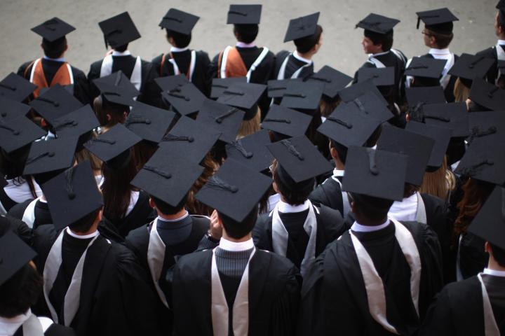 Graduates in black caps and gowns at a ceremony, viewed from behind.