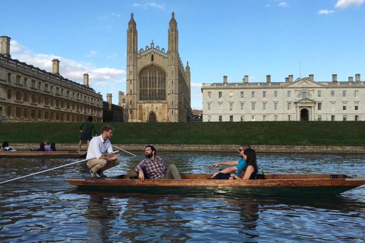 Three people in a wooden boat on a river, historic building in the background.