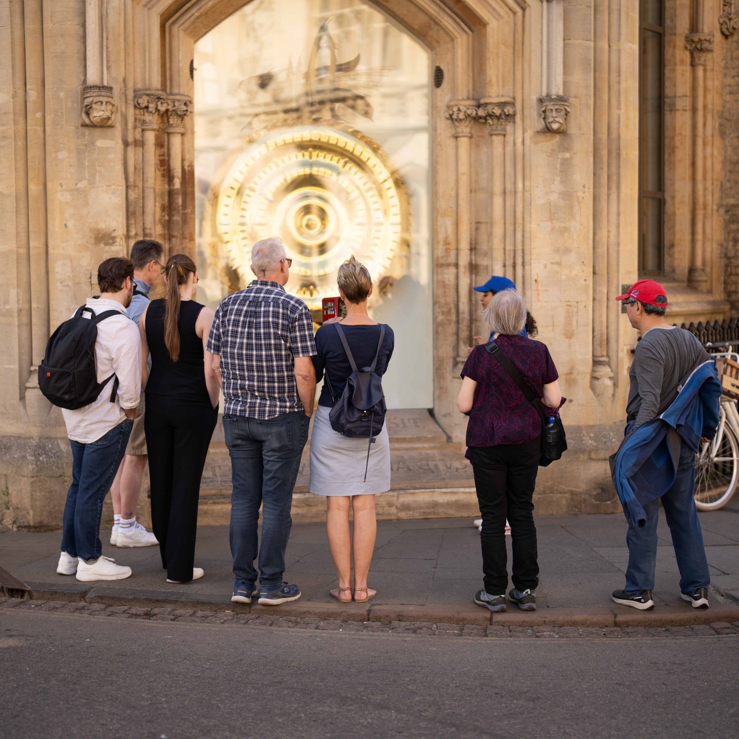 Group of people stand on pavement, looking at a large golden clock in a historic building.