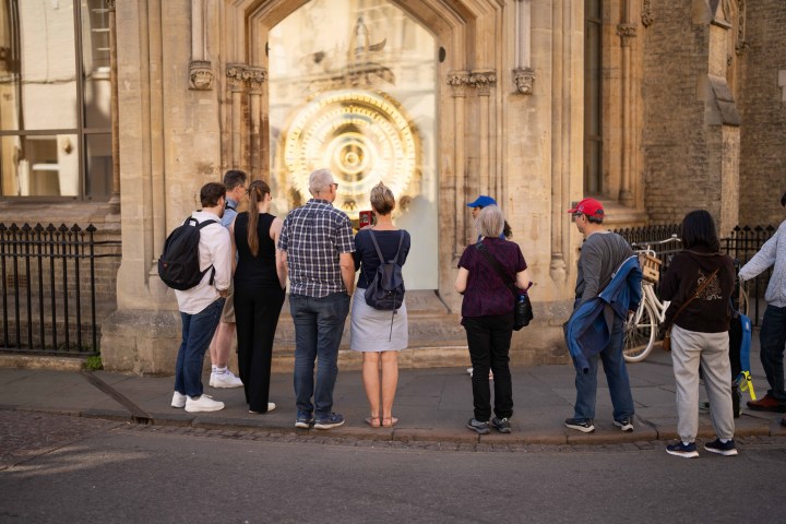 Group of people stand on pavement, looking at a large golden clock in a historic building.