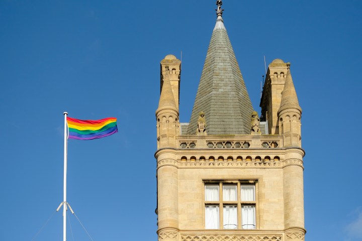 Rainbow flag flying next to a historic building tower under a clear blue sky.