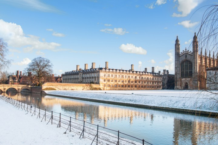 Snowy landscape with historic building, river, and bridge under a blue sky.