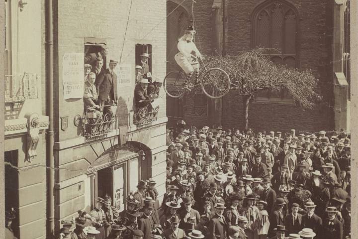 A crowd watches a woman on a bicycle suspended from a building in a historical setting.