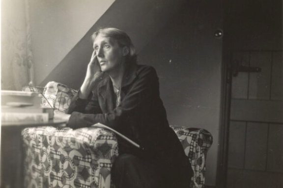 Person sitting on patterned chair in thought, hand on face, next to a stack of books.