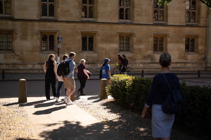 People walking on a path near a historic building with sunlight and shadows.