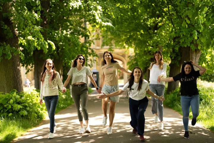 Six women joyfully walking on a tree-lined path in sunlight.