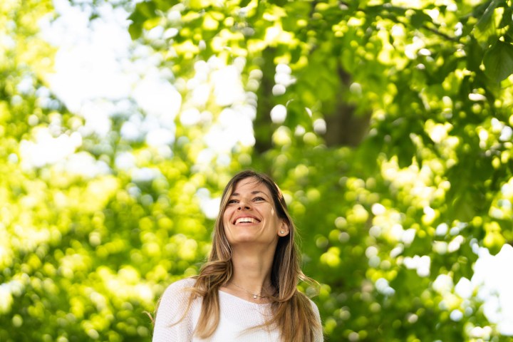 Smiling woman in white top under bright green trees.